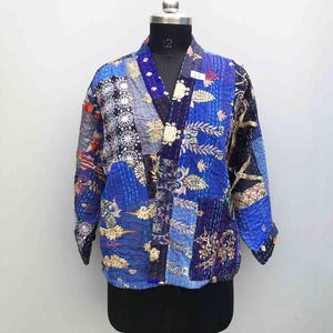 Op de afbeelding: Een jas met lange mouwen met een patchworkontwerp van diverse blauwe stoffen met ingewikkelde gouden bloemenborduursels en witte cirkelvormige patronen. Het kledingstuk heeft een V-hals, een opening aan de voorzijde en zichtbare kantha-stiksels.