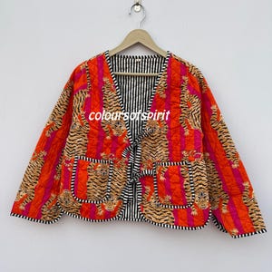 以下が含まれることがあります： オレンジとピンクの虎柄のキルティングジャケット。黒と白のストライプの裏地とトリム、2つのポケット、タイクロージャーが付いています。「coloursofspirit」の文字が見えます。