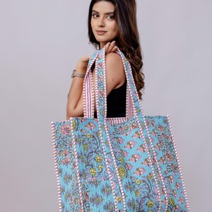 May include: A large quilted tote bag featuring a light blue base with a repeating floral pattern in shades of pink, green, and brown. The bag has two sturdy shoulder straps and is finished with white and pink striped fabric trim along the edges.