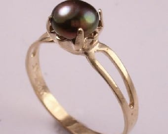 Puede incluir: Anillo de oro con una perla oscura e iridiscente. La perla está sujeta por cuatro garras. El anillo tiene una banda de caña dividida. El fondo es de un color suave y neutro. Una pieza de joyería clásica.
