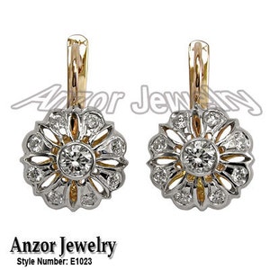 Pode incluir: Um par de brincos em forma de flor com design dourado e prateado. Cada brinco apresenta um diamante central rodeado por diamantes menores e um fecho dourado. Joalharia Anzor, modelo E1023.