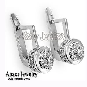 Può includere: Un paio di orecchini argentati con un design rotondo e strutturato che racchiude un gruppo di piccole pietre scintillanti. Gli orecchini hanno una chiusura curva a forma di gancio. Il testo "Anzor Jewelry" e il numero di stile "E1016" sono visibili.