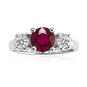 May include: A silver ring featuring a large, round, deep red ruby center stone, flanked by two smaller, round, clear diamond side stones. The ring has a classic three-stone design, with the gemstones set in a prong setting.