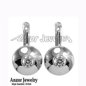 Peut inclure: Une paire de boucles d'oreilles argentées avec un design rond et poli. Chaque boucle d'oreille présente une pierre précieuse centrale et étincelante. Fermeture à levier. La marque "Anzor Jewelry" et le numéro de modèle "E1034" sont visibles.