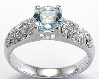 May include: A silver ring featuring a round, light blue gemstone. The band has intricate filigree detailing with small accent stones. The ring is set against a white background, highlighting its elegant design.