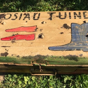 May include: Vintage trunk with the name "JOSIAU TUINER" in black lettering. The trunk features painted illustrations of red shoes and a blue boot. The trunk has a weathered, distressed appearance with visible rust and wear.