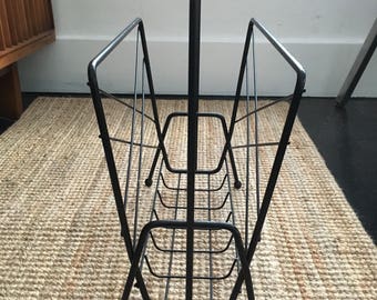 May include: Black metal magazine rack with a handle. The rack has a wireframe design with a rectangular base and angled sides. The rack is designed to hold magazines and newspapers. The rack is on a woven rug.
