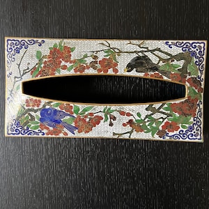 Könnte beinhalten: Ein rechteckiger Gewebeschachtel-Deckel aus Messing mit weißem Emaille-Hintergrund und einem floralen Design. Das Design zeigt rote und rosa Blüten, grüne Blätter und zwei Vögel, einen schwarzen und einen blauen.