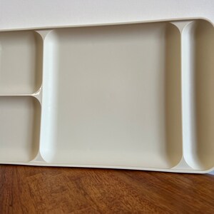 Op de afbeelding: Een rechthoekige, crèmekleurige plastic dienblad met afgeronde hoeken en vier afzonderlijke compartimenten. Het dienblad is ontworpen met een groot centraal gebied en drie kleinere, verdeelde secties, ideaal voor het organiseren van items. Het dienblad staat op een houten oppervlak.