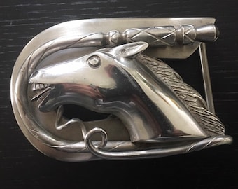 May include: A silver belt buckle with a horse head design. The horse's mouth is open and its mane is detailed. The buckle is shaped like a horseshoe and has a decorative design.