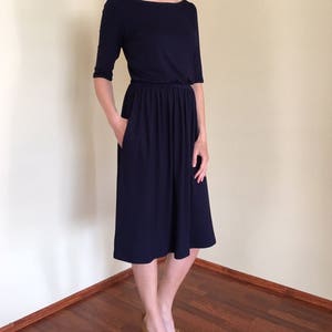 Op de afbeelding: Een donkerblauwe midi-jurk met een boot halslijn en 3/4 mouwen. De jurk heeft een geplooide taille en een licht uitlopende rok.