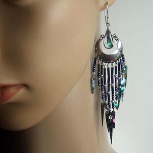 May include: Silver-toned dangle earring with a crescent moon design and dangling iridescent accents. The earring features a teardrop-shaped top and multiple strands of beads and iridescent pieces, creating a layered effect.