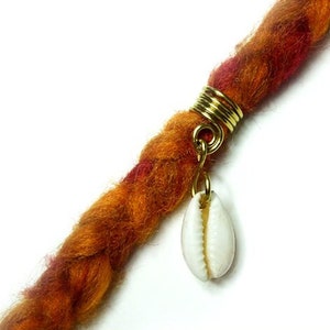 May include: A handmade dreadlock extension in shades of orange, red, and brown, adorned with a gold-colored metal ring and a small white seashell charm. The word "Antonina" is written in cursive.