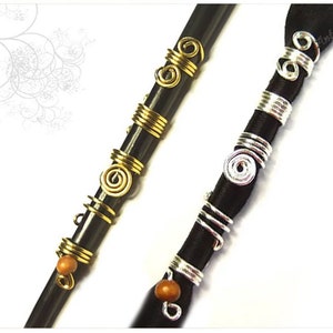 May include: Close-up of two dark hair wraps adorned with decorative metal wire and wooden beads. One wrap features gold-colored wire in spiral and coiled designs. The other wrap has silver-colored wire in similar patterns. The wraps are against a white background.