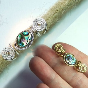 Könnte beinhalten: Zwei Dreadlock-Perlen, eine silberne und eine goldene, mit einem grünen und blauen Abalone-Muschel-Akzent.