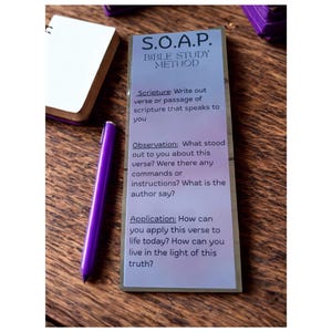 May include: A purple pen rests beside a small white notebook and a card with the text "S.O.A.P. BIBLE STUDY METHOD." The card has instructions for scripture study, including "Scripture," "Observation," and "Application." The background is a wooden surface.