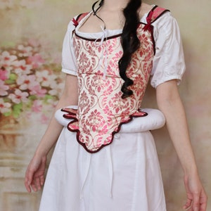 May include: A white and pink brocade corset with a scalloped hem and a burgundy trim. The corset is worn over a white dress.