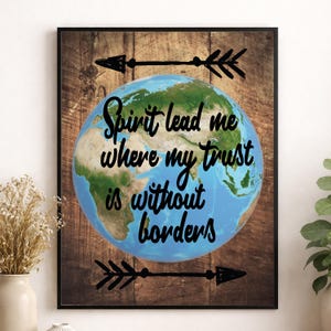 以下が含まれることがあります： 「Spirit lead me where my trust is without borders」のテキストが入った地球のイラストが描かれた額入りのプリント。地球は木製の背景に配置され、テキストの上と下に黒い矢印のデザインが施されています。