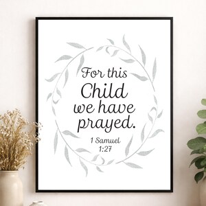 以下が含まれることがあります： 「For this Child we have prayed」というテキストがエレガントな筆記体で書かれた額入りのプリント。銀灰色の花のリースで囲まれています。「1 Samuel 1:27」という聖書の引用が下にあります。白い背景に飾られたアートワークです。