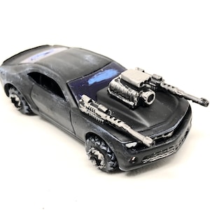 May include: A black toy car with a large engine and two machine guns mounted on the hood. The car has a weathered look and is on a white background.