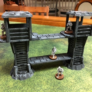 May include: A gray plastic model of a two-level tower with a bridge connecting the two levels. The tower has a small, open room on the top level. There are four small, gray plastic figures standing on the bridge and the top level of the tower.