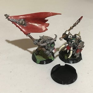 May include: Two miniature fantasy figures, one holding a red banner with a skull, the other with a spiked weapon and shield. The figures are on black bases, with a separate black base in the foreground. The figures are painted in shades of green, gray, and red.
