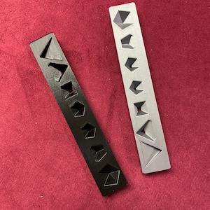 May include: Two black and gray plastic rulers with geometric shapes cut out. The rulers are on a red surface.