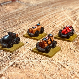 May include: Miniature toy vehicles in various colors, including black, orange, and red. Each vehicle has large wheels and is mounted on a small, square base. The vehicles appear to be part of a tabletop game or hobby.