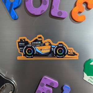 May include: A magnetic orange race car with black wheels and colourful accents. The car has the words "android", "alterx analytics", "vuse", and "now" on it. The car is on a silver refrigerator with the letters "POLE" and "FI" in purple and blue.