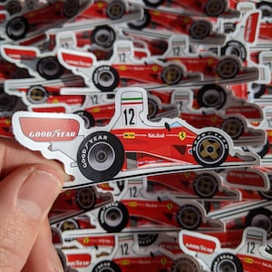May include: A red and white sticker of a Formula One race car with the number 12 and the name "Niki Lauda" on it. The car is on a Goodyear tyre and has the Italian flag on the side. The sticker is surrounded by other similar stickers.