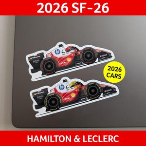 Könnte beinhalten: Zwei Aufkleber eines roten, weißen und schwarzen Rennwagens mit dem Text "2026 SF-26" oben und "2026 CARS" auf einem gelben Kreis. Der untere Teil des Bildes lautet "HAMILTON & LECLERC". Die Aufkleber befinden sich auf einer grauen Oberfläche.