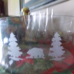 May include: Clear glass cylinder vase with etched white silhouettes of pine trees and a bear. The vase has a wide, open top and a slightly curved shape. The design suggests a nature or woodland theme.