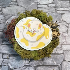 Könnte beinhalten: Eine weiße und gelbe Schlange mit roten Augen, die sich zu einem Kreis zusammenrollt. Die Schlange befindet sich auf einem grünen Moos-Hintergrund.