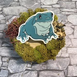May include: A teal frog sticker with a white outline sits atop a bed of green moss and dried foliage. The frog has a smiling expression and is set against a gray stone background. The sticker is a whimsical design.