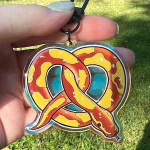 Pode incluir: Um chaveiro de plástico transparente com uma cobra vermelha e amarela em forma de pretzel sobre um fundo azul e branco.