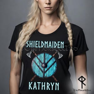 Könnte beinhalten: Schwarzes T-Shirt mit einem blauen Schild und gekreuzten Äxten. Der Text "Shieldmaiden" und "Kathryn" ist in Weiß auf das Shirt gedruckt.