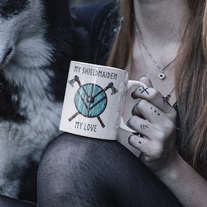 May include: A woman with long braided hair and intricate makeup sits on stone steps with a black and white husky dog. She is holding a white mug with the text "My Shieldmaiden, My Love" and a blue shield with axes on it.