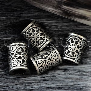 May include: Five silver metal hair beads with intricate designs, including a snowflake pattern, on a black and white furry surface. The Norse rune for protection, Algiz, is visible in the bottom right corner.