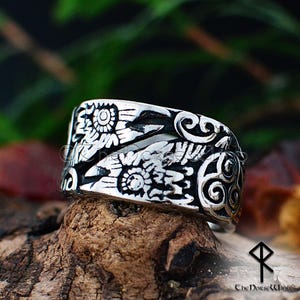 May include: A silver ring with a detailed design of two ravens with their wings spread. The ravens are facing each other and their wings are intertwined. The ring is set against a background of green foliage.