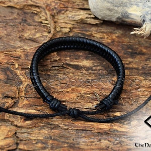 May include: Black leather bracelet with a braided design and adjustable cord closure. The bracelet is tied in a knot and is lying on a wooden surface.