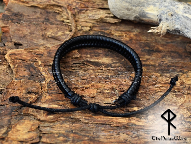 May include: Black leather bracelet with a braided design and adjustable cord closure. The bracelet is tied in a knot and is lying on a wooden surface.