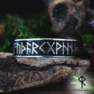 May include: A black and silver stainless steel bracelet with a band of black enamel and raised silver runes. The runes are the Elder Futhark alphabet, which is an ancient Germanic alphabet.