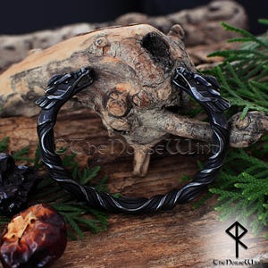 May include: A dark gray metal bracelet with dragon head terminals. The bracelet has a twisted rope design and is displayed on a wooden surface with greenery. The Norse Wind logo is visible.