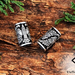 May include: Two silver-toned metal hair beads with a detailed design of a bird with outstretched wings. The beads are on a wooden surface.