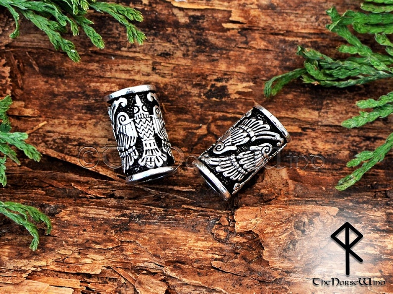 May include: Two silver-toned metal hair beads with a detailed design of a bird with outstretched wings. The beads are on a wooden surface.