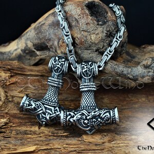 May include: A silver Mjolnir pendant necklace with intricate knotwork and a Viking design. The necklace is on a silver chain and is resting on a piece of wood. The pendant is a symbol of Thor, the Norse god of thunder.