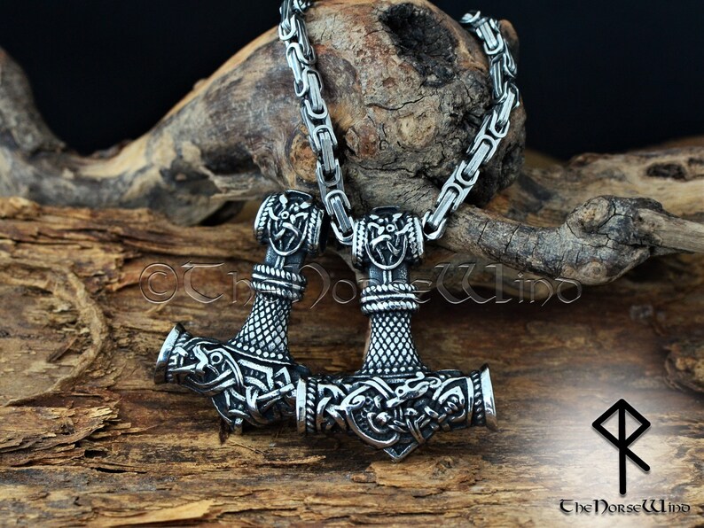 May include: A silver Mjolnir pendant necklace with intricate knotwork and a Viking design. The necklace is on a silver chain and is resting on a piece of wood. The pendant is a symbol of Thor, the Norse god of thunder.
