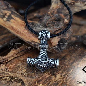 May include: A silver Mjolnir pendant necklace with intricate details, including a braided leather cord. The pendant features a detailed design of the hammer head with a knotwork pattern and a small shield on the top. The pendant is on a wooden surface.