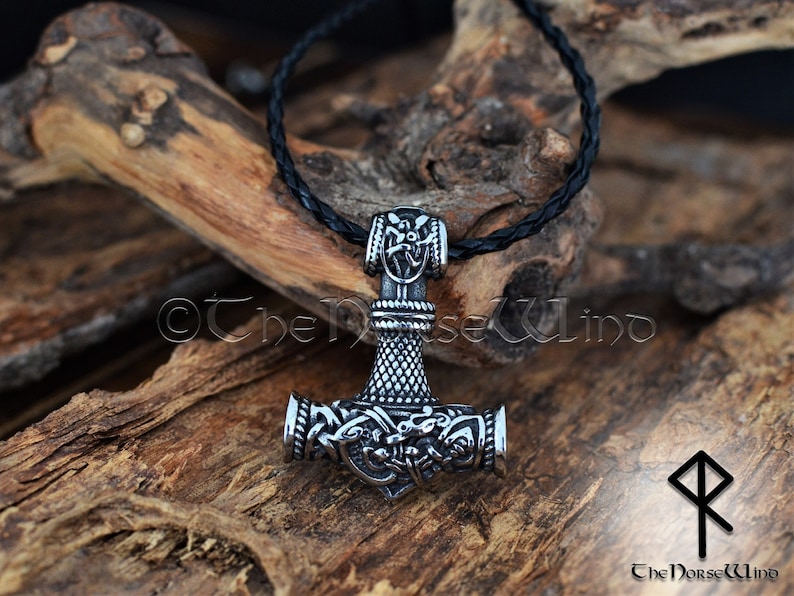 May include: A silver Mjolnir pendant necklace with intricate details, including a braided leather cord. The pendant features a detailed design of the hammer head with a knotwork pattern and a small shield on the top. The pendant is on a wooden surface.