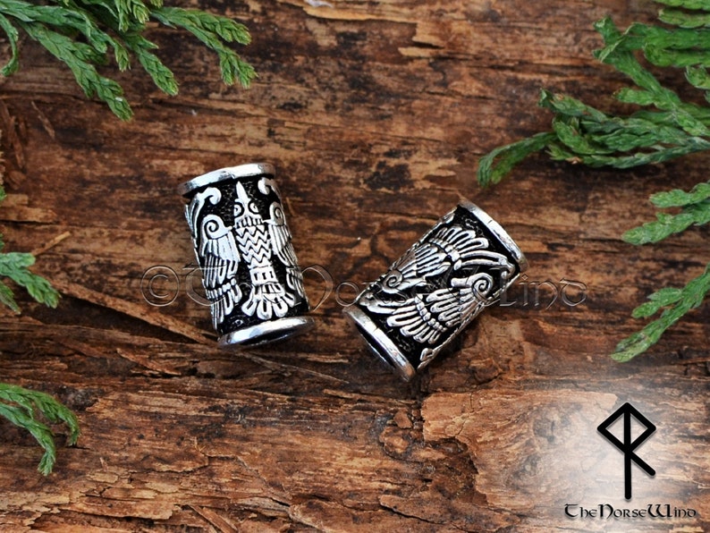 May include: Two silver metal hair beads with a detailed design of a double-headed eagle. The beads are on a wooden surface.